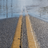 Standing water covers a paved road, partially obscuring its double yellow center lines.
