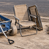 A damaged blue beach cart, two worn beige folding chairs, and a separate pile of branches sit on concrete beside a curb and asphalt street.