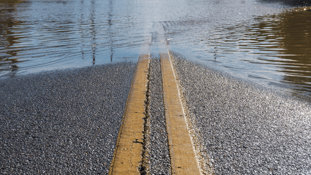 Standing water covers a paved road, partially obscuring its double yellow center lines.